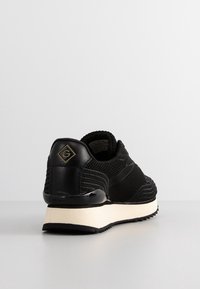 Czarny sneaker z teksturowanym dzianinowym materiałem i skórzaną piętą, białą podeszwą oraz złotym logo "G" z tyłu, zaprezentowany pod kątem z tyłu w trzech ćwiartkach.