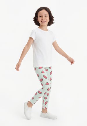 Mosolygó gyermek fehér pólóban, világos színű leggingsben görögdinnyeszeletekkel, és fehér bebújós cipőben, karjait kissé kinyújtva áll.