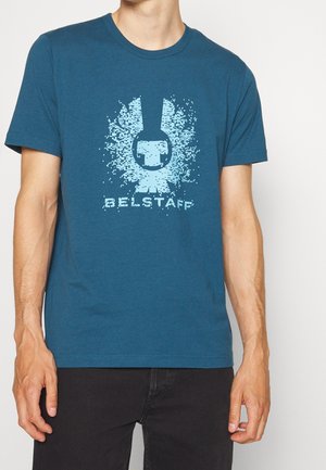 Mężczyzna noszący niebieską koszulkę z jasnoniebieskim zdeformowanym logo i napisem Belstaff na piersi, w połączeniu z czarnymi spodniami.