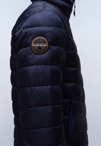 Granatowa kurtka puchowa z pikowanym wzorem, błyszczącą fakturą i haftowaną naszywką na rękawie. Posiada boczny zamek i wysoką kołnierz.