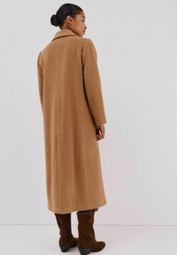 Lungo cappotto di lana marrone con colletto a punte e silhouette dritta. La texture del tessuto appare morbida. Abbinato a stivali marroni scuri fino al ginocchio.