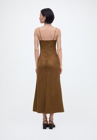 Γυναίκα που στέκεται με την πλάτη γυρισμένη, φορώντας ένα καφέ midi φόρεμα με λεπτές τιράντες και μαύρα σανδάλια με χοντρό τακούνι, σε λευκό απλό φόντο.