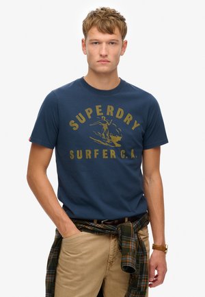 Camiseta de algodón azul marino con un gráfico dorado de "Superdry Surfer C.A." y un diseño de surfista. Pantalones marrón claro con una camisa de cuadros atada a la cintura.