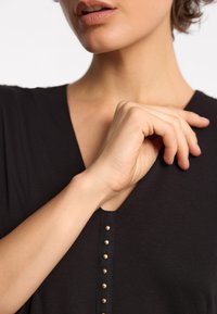 Kobieta w czarnym topie delikatnie dotykająca swojego dekoltu prawą ręką, częściowo odsłaniająca usta i brodę na jednolitym tle.