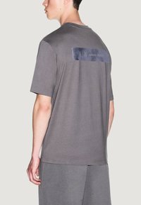 Camiseta de algodón gris con mangas cortas, cuello redondo acanalado y un parche rectangular azul texturizado con el logo en la parte posterior.