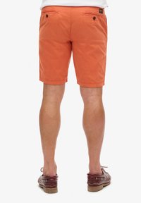 Pomarańczowe bawełniane szorty o długości do połowy uda, z dwiema tylnymi kieszeniami zapinanymi na guziki. Szorty mają gładką teksturę i regularny krój.