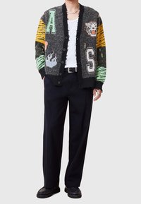 Pletený cardigan s multicolorovými motivy zvířat a plamenů, knoflíky vpředu a texturovaným povrchem. Nositý přes bílou košili a černé kalhoty.