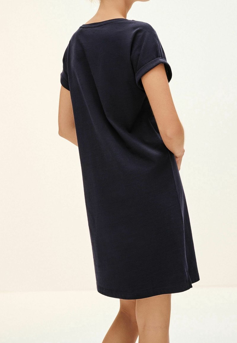 Femme portant une robe bleue marine à manches courtes et longueur genou avec les revers retroussés, debout les mains dans les poches, tournée de dos à la caméra.