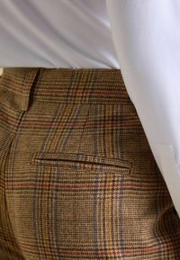 Rjave hlače s karo vzorcem in teksturirano tkanino. Vzorec vključuje modre in rdeče črte, na zadnji strani imajo žep in so krojene po meri. Nošene so z belo srajco.