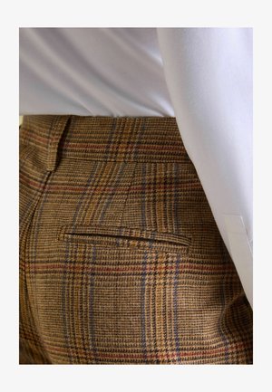 Brązowe spodnie w kratę z teksturowanej tkaniny. Zawierają niebieskie i czerwone linie, tylną kieszeń oraz dopasowany krój. Noszone z białą koszulą.