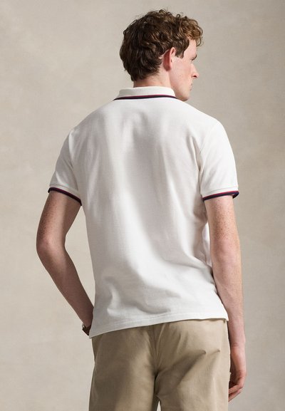 Göndör hajú férfi, fehér rövid ujjú galléros ingben, tengerészkék és piros szegéllyel, bézs nadrággal, háttal áll a kamerának.