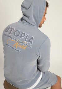 Felpa con cappuccio in tessuto grigio chiaro con testo ricamato: "UTOPIA ILLUSIONED ALTERNATIVE WORLD" in colori misti, con polsini a costine.