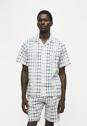 Mand iført matchende kortærmet skjorte med knapper og shorts med et blåt og hvidt geometrisk mønster, der står foran en ensfarvet baggrund.
