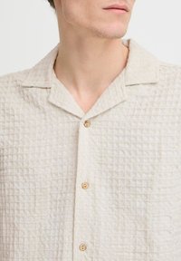 Camisa de manga corta beige con un patrón texturizado en forma de cuadrícula, que presenta un cuello y botones de madera, fabricada con un tejido ligero.