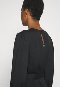 Personne aux cheveux courts et bouclés portant une robe noire à manches longues avec une petite ouverture en goutte et une fermeture par bouton au niveau de la nuque dans le dos.