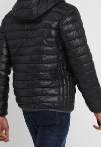 Svart puffjacka med horisontell sömnad, huva och glänsande yta. Har en figursydd design och ribbad fåll, tillsammans med blå jeans.