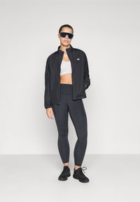 Juodas lengvas džemperis su užtrauktuku priekyje, prigludusi balta trikotažo palaidinė, aukštu juosmeniu juodos leggings ir juodi sportiniai bateliai. Dideli tamsūs akiniai nuo saulės.