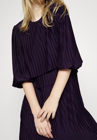 Djupt lila faltad klänning med ett lagerdesign och korta puffärmar. Slät textur och samlad halsringning. Modellens händer korsade fram.