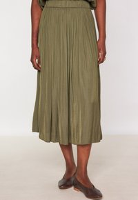 Gonna midi plissettata verde oliva con vita elastica, realizzata in tessuto leggero, abbinata a scarpe scivolate marrone scuro.