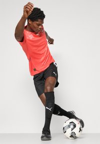 Rød Puma fodboldtrøje med subtile detaljer, sorte shorts, knæhøje sorte sokker, sorte fodboldsko og en hvid fodbold med sorte accenter.