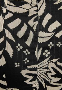 Tissu noir avec un imprimé botanique crème audacieux, comportant des feuilles et des fleurs. Surface texturée avec de légères lignes verticales. Détail de fermeture éclair à l'arrière.