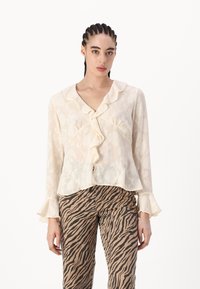 Cremefarbene Bluse mit Rüschensaum an Kragen und Ärmeln, verziert mit einem floralen Muster. Kombiniert mit gestreiften Hosen in Braun und Beige.
