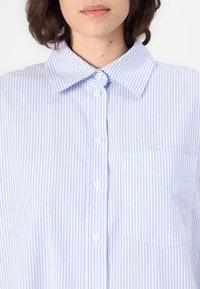 Camisa de botão listrada em azul claro e branco, apresentando um colarinho clássico, bolso frontal e tecido texturizado. Os botões são brancos e proeminentes.