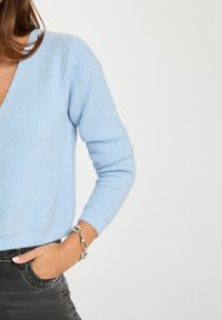 Helles blaues Strickpullover mit V-Ausschnitt, gerippter Textur, langen Ärmeln und dezenten Glitzerpartikeln, getragen mit einem grauen Rock und einem silbernen Armband.