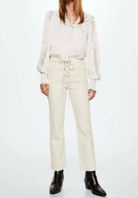 Blouse blanche texturée avec col à volants et patte de boutonnage ; jeans crème clair taille haute avec lacets sur le devant ; bottines noires.