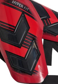 Rote und schwarze Schienbeinschoner mit einem eleganten Design, das ein geometrisches Muster und den Text "HYPER FIT" zeigt. Enthält einen Riemen für sicheren Halt.