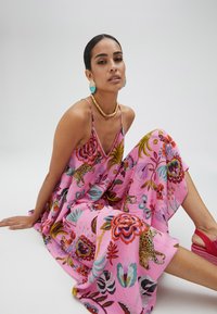 Robe maxi florale rose avec des motifs animaliers et botaniques vibrants, bretelles spaghetti réglables et fentes sur les côtés pour le mouvement.