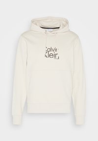 Crème hoodie van zacht materiaal, met een groot zwart "Calvin Klein" logo op de voorkant en een capuchon met trekkoord.