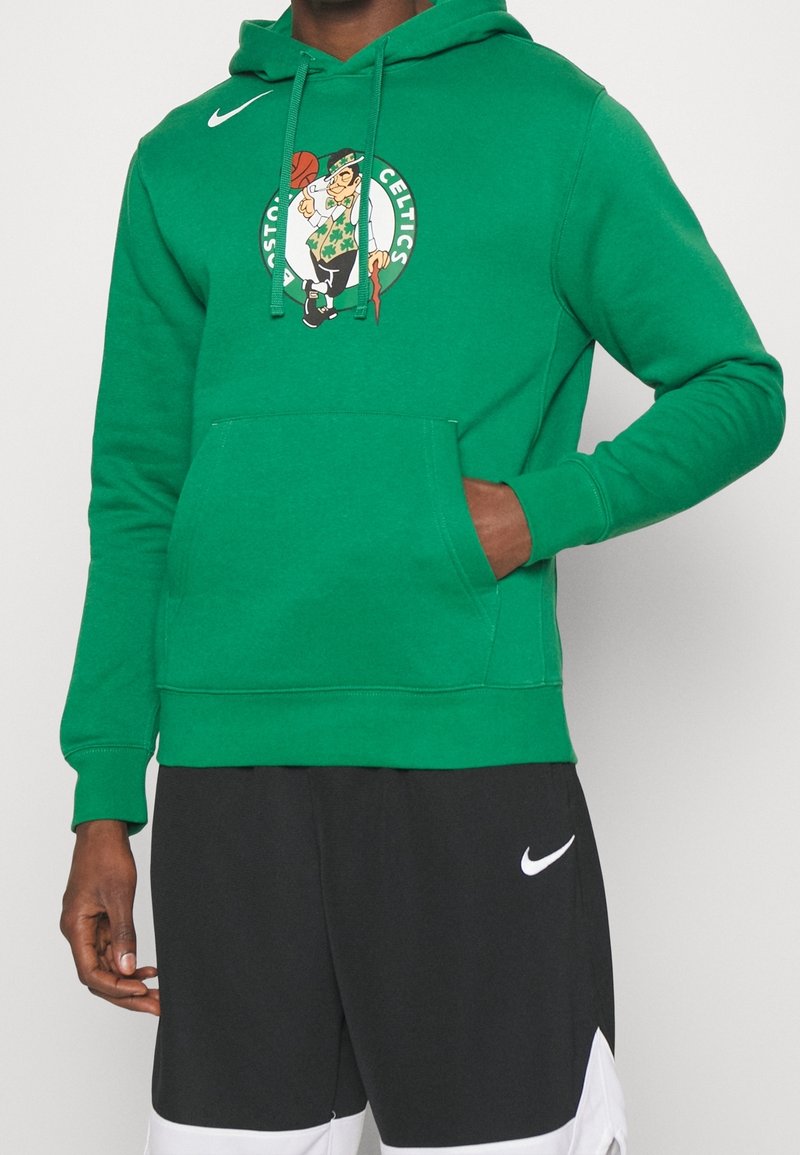 Personne portant un sweatshirt vert des Boston Celtics avec le logo et un short noir Nike, une main dans la poche du sweatshirt, sur fond uni.
