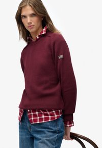 Burgundy knitted sweater with ribbed neckline and cuffs, layered over a red and white checkered shirt. Denim jeans complete the outfit.