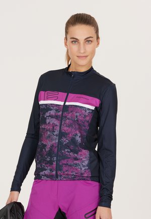 Långärmad cykeljacka i marinblått och lila, med ett texturerat mönster, framdragkedja och hög krage. Bärs med lila shorts.