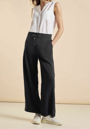 Femme portant un haut sans manches blanc, un pantalon large noir avec taille à cordon, et des baskets blanches, se tenant avec les mains dans les poches.