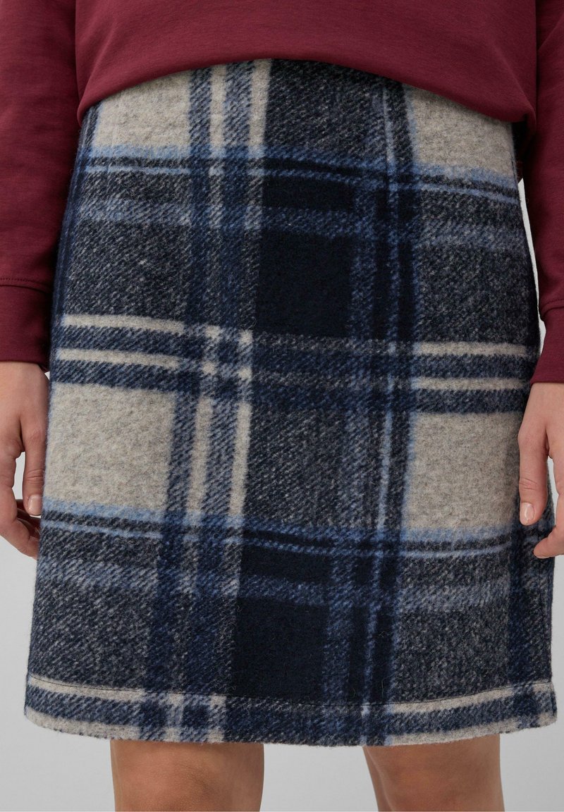 Jupe mi-longue à motif écossais bleu marine, bleu et beige, portée avec un haut à manches longues rouge foncé visible à la taille.