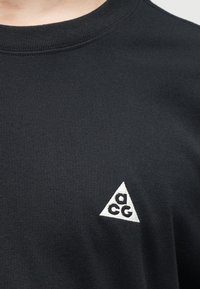 Μαύρο μπλουζάκι με στρογγυλή λαιμόκοψη, με μικρό λευκό τριγωνικό λογότυπο που φέρει τα γράμματα "a", "c" και "g" στην αριστερή πλευρά του στήθους.