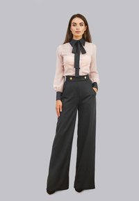 Jasnoróżowa bluzka z czarnym kołnierzykiem i muszką, połączona z wysokim czarnym spodniami o szerokich nogawkach z złotymi guzikami i kieszeniami.