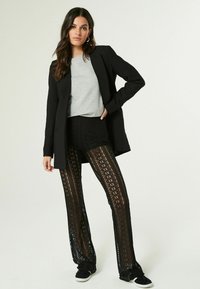 Blazer nero sovrapposto a una camicia grigia, abbinato a pantaloni neri in pizzo a vita alta con motivi floreali. Sneakers nere completano l'outfit.