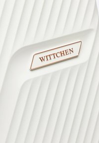 Biała, teksturowana walizka o twardej skorupie z pionowymi żłobieniami, z metalową płytką z logo w kolorze złotym "WITTCHEN".