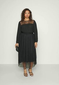 Fekete, hosszú ujjú ruha áttetsző csipke felsőrésszel és pliszírozott szoknyával, szűkített derékkal. Kiegészítői között szerepel egy kis fekete táska és pántos szandál.