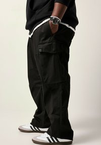 Schwarze Cargo-Hosen mit Seitentaschen, kombiniert mit einem weißen geschichteten Hemd und grau-weißen Turnschuhen mit schwarzen Streifen.