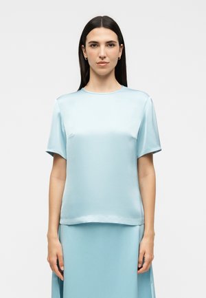 Kobieta o długich ciemnych włosach, ubrana w jasnoniebieski satynowy top z krótkim rękawem i pasującą spódnicę, stojąca na tle jednolitego jasnego tła.