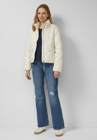 Giacca trapuntata color crema con colletto alla coreana, zip frontale e due tasche, abbinata a top scuro e jeans flare blu slavati con effetto consumato. Sneakers bianche.