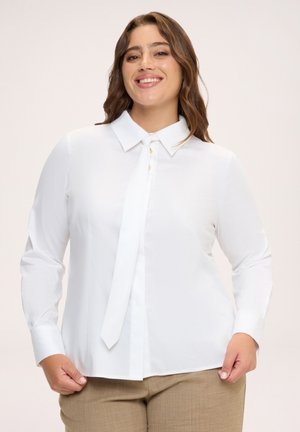 Smilende kvinde med brunt hår iført en hvid bluse med lange ærmer og slips samt beige bukser mod en ensfarvet lys baggrund.
