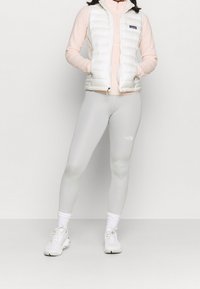 Gilet en duvet blanc sur un haut à manches longues rose pâle, associé à des leggings gris et des baskets blanches. La tenue présente un design épuré et minimaliste.