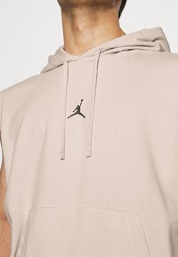 Beige sleeveless hoodie with a front pocket and drawstring hood. Features a black Jumpman logo on the chest. Soft, smooth fabric texture.