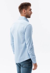 Camisa polo de manga larga de color azul claro con una textura suave, puños abotonados y un cuello clásico, combinada con unos vaqueros.