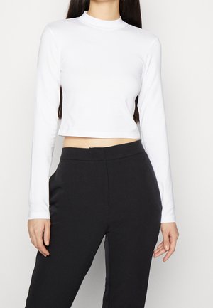Kvinde iført en ensfarvet hvid langærmet cropped top og højtaljede sorte bukser, stående mod en hvid baggrund.